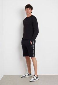 Camisa negra de manga larga con el logo "GUESS" en relieve, combinada con pantalones cortos negros que tienen una franja lateral. Zapatillas negras y blancas completan el look.
