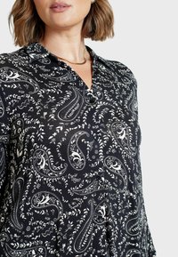 Zwarte blouse met een paisley- en bloemenpatroon in het wit, met een kraag, knoopsluiting aan de voorkant en een soepele stoffen textuur.