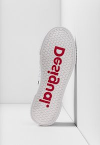 Valkoinen lenkkaripohja, jossa on teksturoitu pinta; siinä on suuri punainen "Desigual"-logo. Pyöreä muoto ja joustava materiaali, joka soveltuu rentoon käyttöön.
