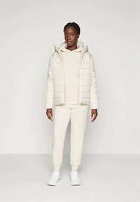Pufferjakke i lys beige med horisontal quilting, brukt over en matchende hettegenser og joggebukser. Hvite sneakers fullfører antrekket.