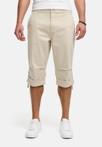 Shorts beige arrivant au genou en mélange de coton, avec des manches roulées et des poches avant, associés à des baskets blanches. Texture lisse.