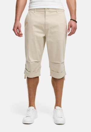 Pantalones cortos beige a la altura de la rodilla, de mezcla de algodón, con puños enrollados y bolsillos frontales, combinados con zapatillas blancas. Textura suave.