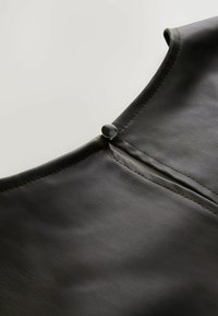 Schwarzes Lederkleidungsstück mit glatter Textur, mit einem runden Halsausschnitt und einem Reißverschluss, der durch ein kleines schwarzes Knopfdetail hervorgehoben wird.