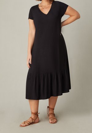 Vrouw draagt een zwarte midi-jurk met korte mouwen en een gerimpelde zoom en bruine platte vetersandalen, staat met één hand op de heup.
