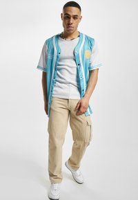 Cămașă albastră deschisă cu mâneci albe, model cu dungi și grafică cu soare; asortată cu pantaloni cargo bej și adidași albi.