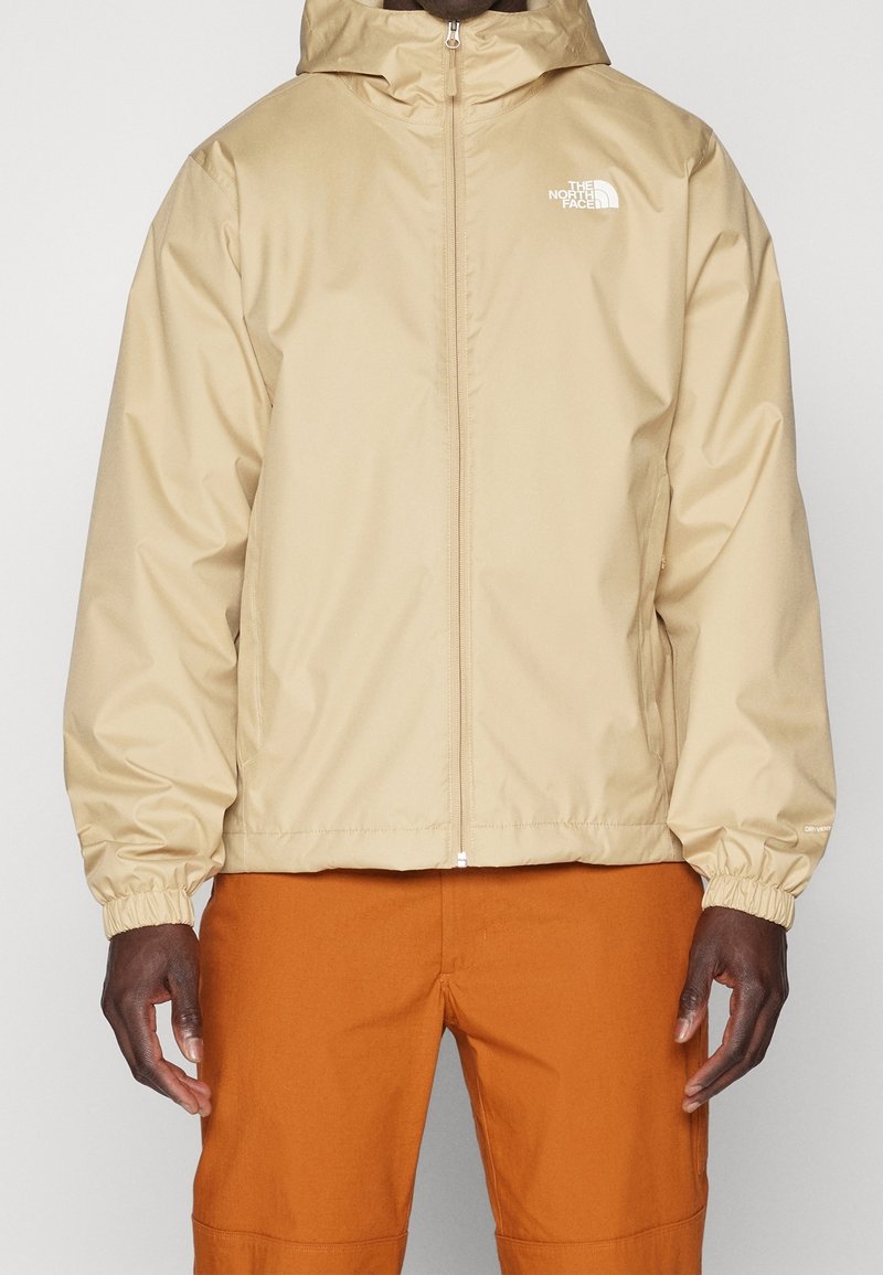 Beige zip-up jacket with hood and elastic cuffs worn with orange pants, featuring The North Face logo on the chest.
