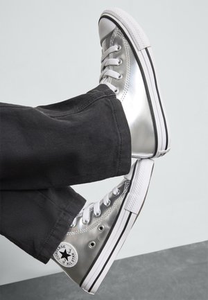 Baskets montantes argentées en matériau brillant, avec des lacets blancs, une semelle noire et blanche, et un patch logo noir sur le côté.