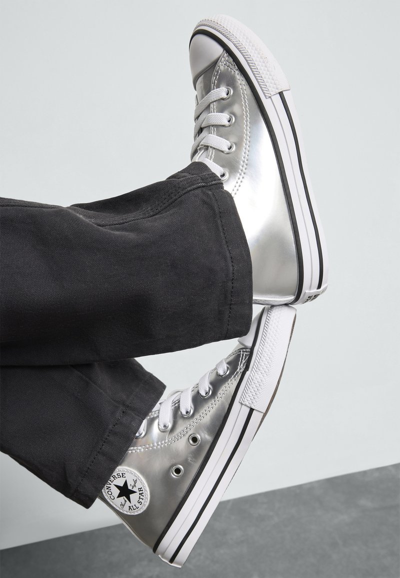 Baskets montantes argentées en matériau brillant, avec des lacets blancs, une semelle noire et blanche, et un patch logo noir sur le côté.
