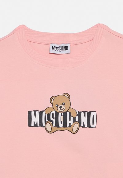Ροζ παιδικό πουκάμισο με στρογγυλή λαιμόκοψη, με ένα αρκουδάκι που κρατάει ασπρόμαυρα γράμματα που σχηματίζουν τη λέξη «Moschino» στο μπροστινό μέρος.