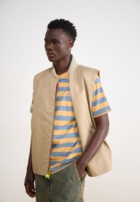 Chaleco sin mangas beige con cuello verde sobre una camisa a rayas azules y amarillas. Pantalones de mezclilla con ligera textura, cremallera visible y detalles de acento.