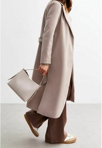 Beige lång kappa med vida slag, kombinerad med bruna vida byxor och vita sneakers. Bär en ljusgrå bucket bag med gulddetaljer.