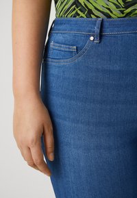 Jeans de mezclilla azul con cintura alta, que presentan un bolsillo lateral con costura visible y una textura suave. Hay una mano descansando cerca.