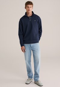 Tengerészkék pulóver félcipzáros gallérral, csíkos ing fölött viselve, világoskék farmerrel és szürke slip-on cipőkkel párosítva. Kényelmes fazon.