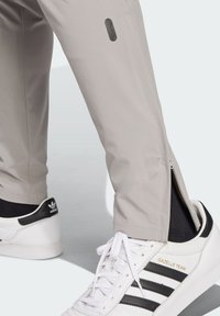 Pantalones deportivos de un gris claro con un detalle de cremallera en el tobillo, con una textura de tela suave. Combinados con zapatillas blancas con acentos en negro.