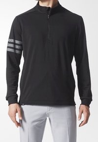Pullover negro de mangas largas y cremallera de un cuarto con tres franjas reflectantes en el brazo derecho, llevado con pantalones gris claro.