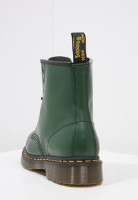 Zielony skórzany botek za kostkę o gładkim wykończeniu, z żółtym szwem, gumową podeszwą oraz czarną i żółtą pętlą oznaczoną napisem "z odbijającymi podeszwami".