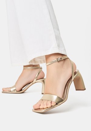 Złote metaliczne sandały na wysokim obcasie z odkrytym palcem, cienkim paskiem wokół kostki i blokowym obcasem. Gładka tekstura z minimalistycznym wzornictwem.
