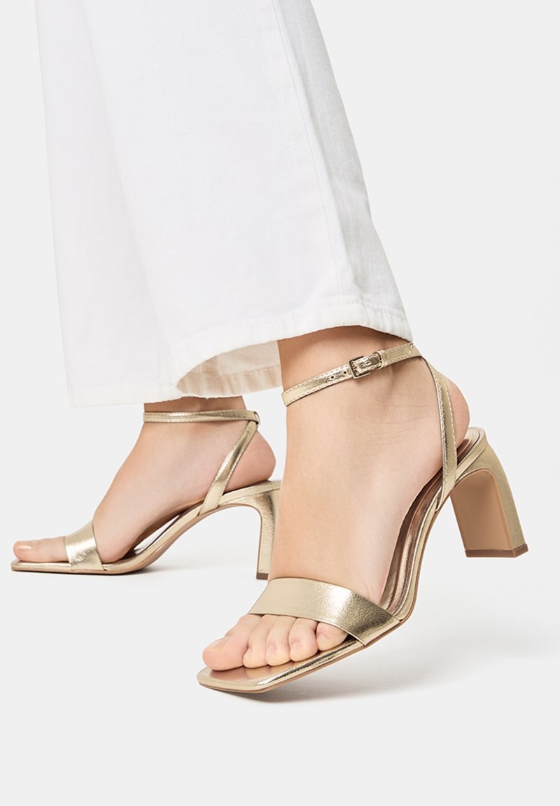 Sandales à talons hauts en métal doré avec bout ouvert, fine sangle de cheville et talon bloc. Texture lisse avec un design minimaliste.