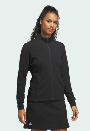 Frau mit lockigem Haar, trägt eine schwarze Reißverschlussjacke und einen passenden schwarzen Rock mit einem kleinen weißen Adidas-Logo nahe dem Saum.