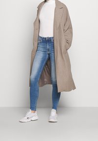 Abrigo largo de lana beige sobre un suéter de cuello alto blanco, combinado con jeans ajustados azules y zapatillas blancas. Textura suave y ajuste relajado.