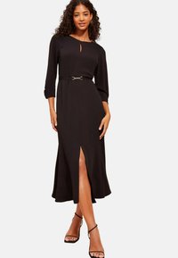 Schwarzes Mittellanges Kleid mit Schlüssel-Ausschnitt, dreiviertel Ärmeln, einem taillierten Gürtel und einem Schlitz vorne. Glattes Material mit minimaler Textur.