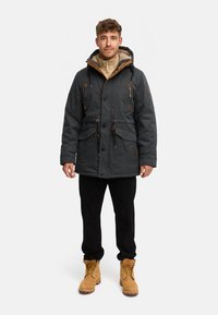 Donkergrijze parka met een capuchon gevoerd met nepbont, bruine accenten, knoopsluitingen en voorzakken. Draagt over een licht beige jas met rits en zwarte broek.