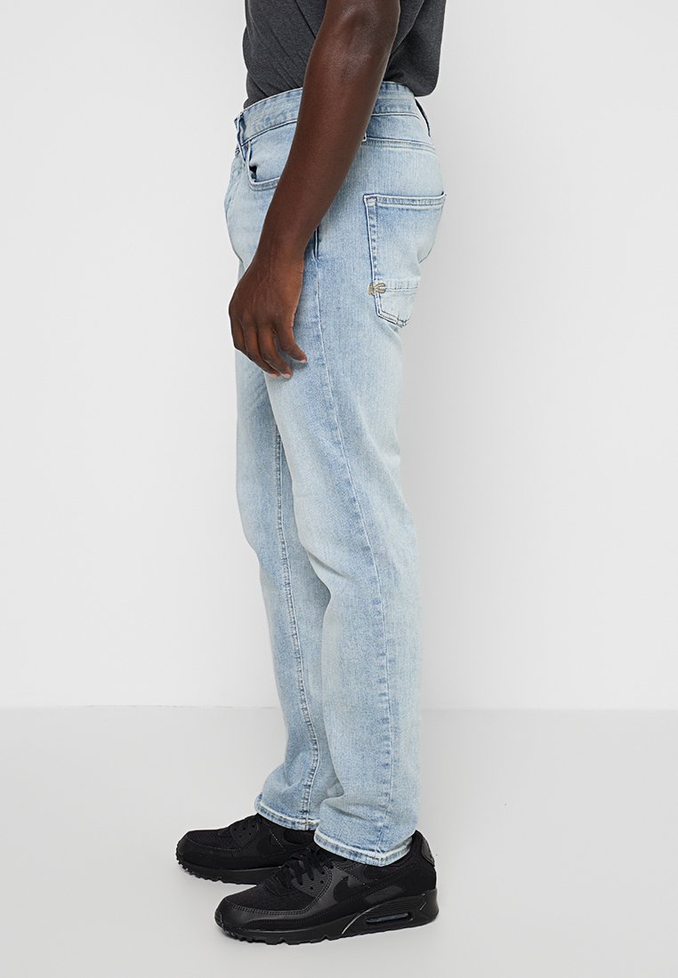 Lichtblauwe denim jeans met een rechte snit, voorzien van een voorzak voor munten, achterzakken en contrasterende stiksels. Gekleed met zwarte sneakers.