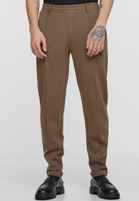Brown sweatpants with a relaxed fit, featuring a smooth texture, elastic waistband, and subtle side piping details. Worn with black shoes.