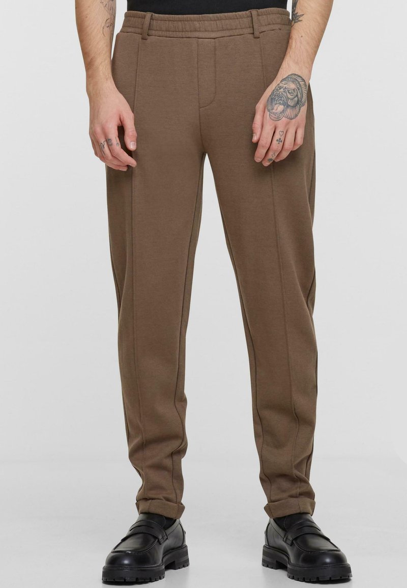 Brown sweatpants with a relaxed fit, featuring a smooth texture, elastic waistband, and subtle side piping details. Worn with black shoes.