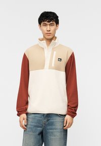 Νέος άνδρας που φοράει μπλουζάκι fleece με χρωματικές μπλοκ σε μπεζ, κρεμ και σκουριά τμήματα, και μπλε τζιν, στέκεται μπροστά σε λευκό φόντο.