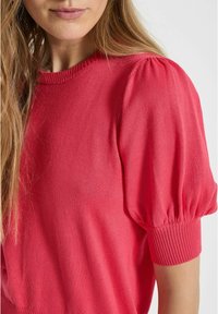 Haut en tricot rouge avec de courtes manches bouffantes, des poignets côtelés et un col ras du cou. Texture lisse avec un léger brillant, mettant en valeur la forme et l'ajustement.