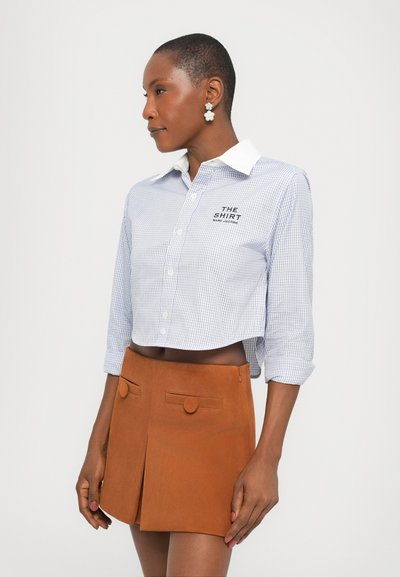 Camicia a quadretti blu e bianca tagliata, con colletto bianco e logo nero, abbinata a una gonna A-line in suede marrone con dettagli circolari.