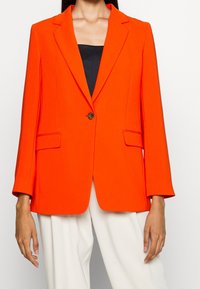 Světlý oranžový blazer se strukturovaným designem, s jedním knoflíkovým zapínáním, klopovými límci a dvěma předními kapsami. Tkanina vypadá hladce a dobře zpracovaně.