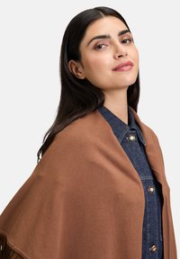 Jeune femme aux longs cheveux noirs portant une chemise en denim et un châle marron, souriant légèrement sur un fond clair neutre.