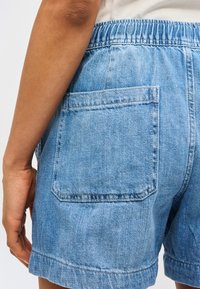 Ανοιχτό μπλε τζιν σορτς με ελαστική μέση και μεγάλη πίσω τσέπη, φορεμένα από ένα άτομο με ορατό χέρι και μπράτσο.