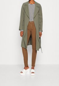 Długi oliwkowozielony płaszcz, czarno-biała pasiasta koszula, brązowe dopasowane spodnie oraz białe sandały na platformie z odkrytymi palcami. Gładkie tekstury i warstwowa konstrukcja.