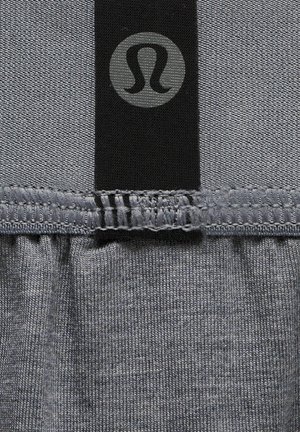 Grå sports top med en sort talje med et cirkulært logo. Stoffet har en glat tekstur med synlige syningsdetaljer.