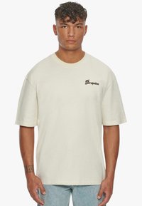 Cremefarvet oversized bomulds T-shirt med korte ærmer, rund hals og et brunt "Drepaize" logo på venstre bryst.