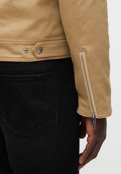 Μπεζ μπουφάν από καμβά με λεία υφή, φερμουάρ στο μανίκι και λεπτομέρεια με πατιλέτα. Φορώντας πάνω από μαύρο τζιν παντελόνι με ορατή πίσω τσέπη.