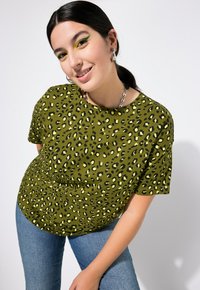 Olivgrünes T-Shirt mit kurzen Ärmeln und einem schwarz-gelben Leopardenmuster. Kombiniert mit hellblauen Jeans. Silberne Kettenhalskette.