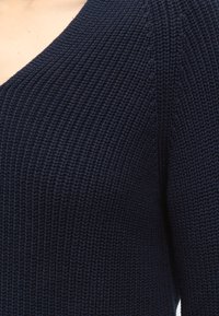 Primo piano di una persona che indossa un maglione lavorato a maglia blu navy scuro con motivo a coste strutturato e scollo a V.