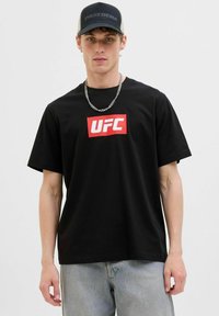 Sort bomuld t-shirt med et fremtrædende rødt UFC-logo på forsiden. Kombineret med lyseblå jeans og en sort/grå kasket. Sølv kædehalskæde.