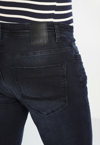 Jeans in denim blu scuro con una toppa in pelle con il logo "CARS" sul retro della vita, caratterizzati da tasche standard e una texture liscia.