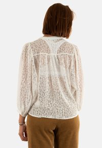 Blusa bianca in pizzo con una vestibilità ampia, maniche a sbuffo e scollatura rotonda. Tessuto traslucido e motivo floreale intricato.