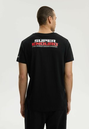 Mężczyzna ubrany w czarny t-shirt z napisem "SUPER ENDURO FIM WORLD CHAMPIONSHIP" drukowanym na plecach na biało i czerwono, stoi tyłem.