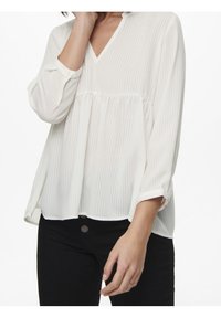 Blusa bianca a maniche lunghe con scollo a V, caratterizzata da un corpetto plissettato e righe orizzontali; abbinata a pantaloni neri.