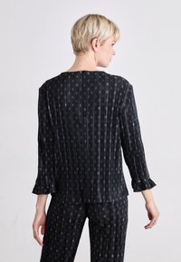 Schwarze, leichte Bluse aus Stoff mit vertikalen Streifen und kleinen runden Mustern. Sie hat einen Drei-Viertel-Ärmel mit Rüschensäumen und einen Knopfverschluss.