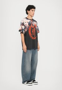 Grafisk T-skjorte med flerfarget abstrakt trykk; svart bunnfarge; oversized design; kombinert med løse, medium-vask denim jeans og sorte sneakers.