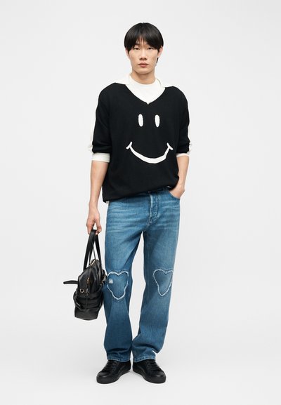 Νέος άνδρας που φοράει μαύρο πουλόβερ με λευκό χαμογελαστό πρόσωπο, μπλε τζιν με μπαλώματα σε σχήμα καρδιάς, μαύρα αθλητικά παπούτσια και κρατάει μια μαύρη τσάντα χειρός.