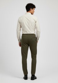 Ljust beige skjorta med krage och mörkgröna smala byxor, med bakficka och elastisk midja, bärs med svarta skor.